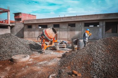 A construction site with a worker wearing safety gear next to a cement mixer and gravel piles.
